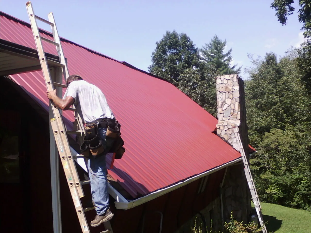 Roofer on a ladder beside a red metal roof and stone chimney — Soffit & Fascia Repair by Roofing Brothers in Blandon